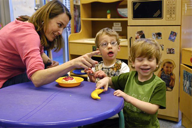 A woman in a pink sweater is pointing at something to two young boys who are sitting at a table. On the table is a plate and a toy banana
